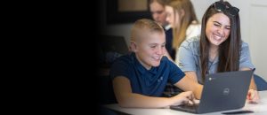 Photo of a smiling student on a laptop being assisted by a teacher