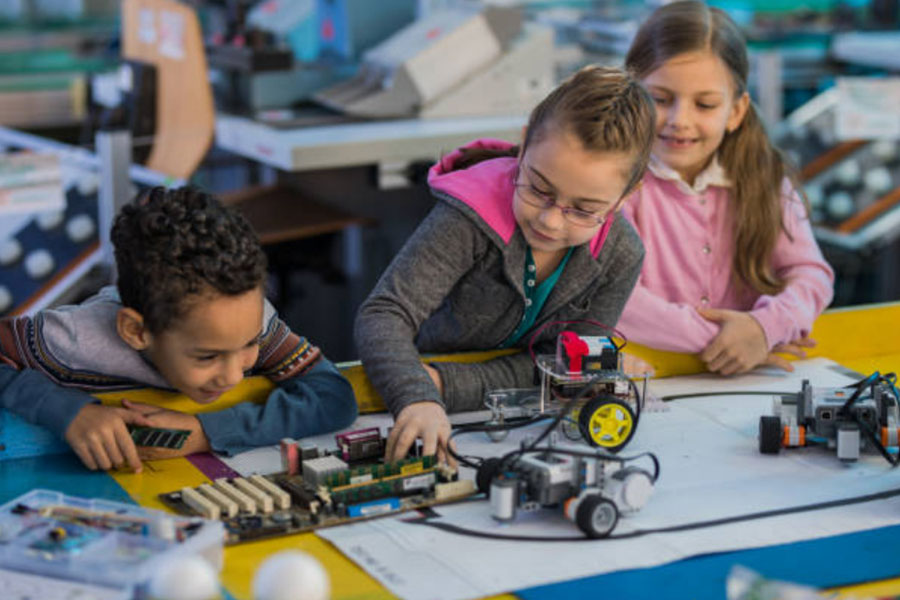 Three kids are playing with circuit boards connected to items with wheels
